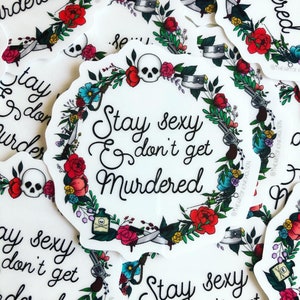 May include: A white sticker with a floral wreath design. The wreath includes a skull, a gun, a knife, a bottle of poison, and a noose. The text inside the wreath reads "Stay sexy & don't get murdered".