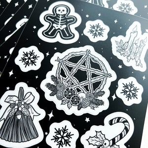 May include: Black and white sticker sheets featuring various holiday-themed illustrations. Designs include a skeleton gingerbread man, snowflakes, a pentagram wreath, a candy cane with skulls, and a broom. The text "the art of jo nicholson" is printed at the bottom.