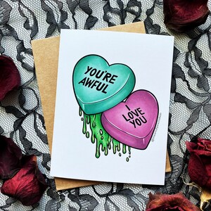 May include: A Valentine's Day card with a green heart-shaped candy that says "You're Awful" and a pink heart-shaped candy that says "I Love You". The candies are dripping green goo.