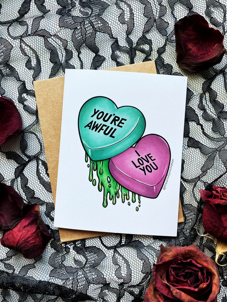 May include: A Valentine's Day card with a green heart-shaped candy that says "You're Awful" and a pink heart-shaped candy that says "I Love You". The candies are dripping green goo.