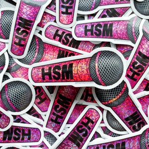 Könnte beinhalten: Ein Stapel pinkfarbener und schwarzer Glitzer-Mikrofon-Aufkleber mit dem Text "HSM" darauf. Die Aufkleber sind in einem zufälligen Muster angeordnet und alle in die gleiche Richtung ausgerichtet.