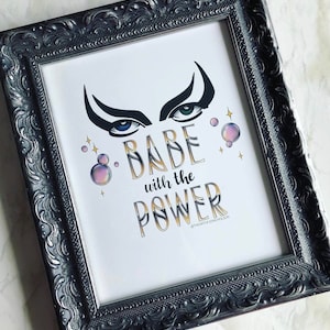 Könnte beinhalten: Ein gerahmter Druck mit weißem Hintergrund und goldener Schrift, die "Babe with the Power" lautet, mit einem Paar stilisierter Augen über dem Text. Die Augen sind blau und grün mit schwarzen Wimpern und Augenbrauen. Um die Augen und den Text herum befinden sich kleine rosa und weiße Blasen und goldene Sterne.