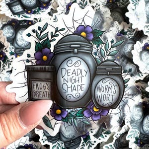 Può includere: Tre barattoli di vetro con coperchi neri ed etichette bianche. Le etichette riportano "Deadly Night Shade", "Worm's Wort" e "Frog's Breath". I barattoli sono circondati da fiori viola e foglie verdi su uno sfondo bianco.