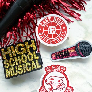 May include: Four stickers featuring popular high school movie themes. The stickers are on a white marble surface. The stickers are: "High School Musical" in gold glitter, "East High Basketball" with a red basketball and white letter E, "East High Wildcats" with a red and white wildcat head, and "HSM" in pink glitter with a microphone.