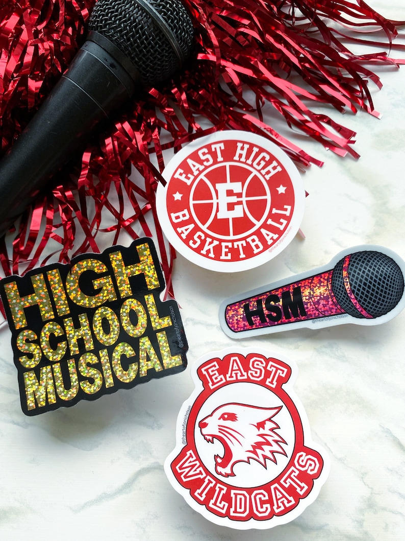 May include: Four stickers featuring popular high school movie themes. The stickers are on a white marble surface. The stickers are: "High School Musical" in gold glitter, "East High Basketball" with a red basketball and white letter E, "East High Wildcats" with a red and white wildcat head, and "HSM" in pink glitter with a microphone.