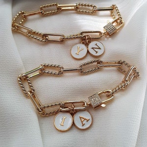 Peut inclure: Deux bracelets en chaîne dorée avec des breloques. Les breloques sont blanches avec des lettres dorées qui épellent les lettres "Y", "N", "I" et "A". Un bracelet a un petit breloque de cadenas avec des strass transparents.