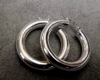 May include: A pair of silver hoop earrings with a thick, rounded band. The earrings are made of sterling silver and have a polished finish.