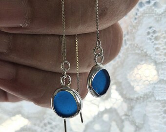 May include: A pair of silver-toned drop earrings with a bright blue, oval-shaped center. The earrings have a chain that hangs down. The background is a soft, white, lacy fabric.