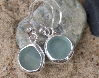 May include: A pair of silver dangle earrings with round, pale blue-green sea glass pendants. The sea glass is set in a silver bezel, and the earrings are displayed on a sandy surface with a gray rock in the background.