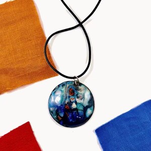 Puede incluir: Un collar con un colgante redondo en un cordón negro. El colgante está hecho de resina y presenta un diseño abstracto azul, verde y blanco en espiral.