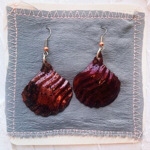 Peut inclure: Une paire de boucles d'oreilles au design unique. Les boucles d'oreilles sont en matériau rouge foncé avec une surface texturée. Les boucles d'oreilles sont suspendues à des crochets argentés.