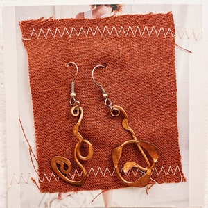 May include: A pair of copper earrings with a unique, abstract design. The earrings are hanging from silver hooks and are displayed on a brown fabric background.