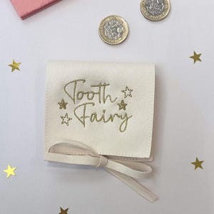 May include: Cream-coloured tooth fairy pouch with gold embroidered text and star accents. The pouch is tied with a matching ribbon. Two gold coins and star-shaped confetti are scattered around the pouch.
