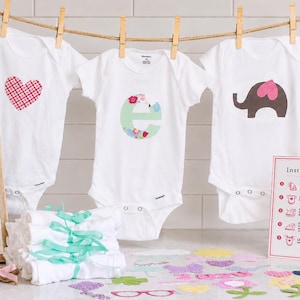 May include: Three white baby bodysuits hanging on a clothesline with wooden clips. One features a red plaid heart, one a floral letter E, and one a brown elephant with a pink heart. Includes a sign reading Please Make A Onesie for the baby.