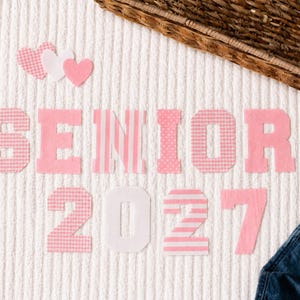 May include: Fabric letters spelling SENIOR and numbers 2027 in various shades of pink, white, and gingham patterns. Three small fabric hearts in pink and white are positioned above the text. The letters and numbers are arranged on a white textured fabric.