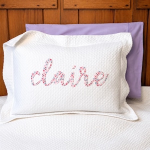 May include: A white quilted pillow sham featuring the name Claire appliqued in a pink and blue floral fabric in a cursive script font. The sham has a wide flange border and is positioned in front of a solid light purple pillowcase.