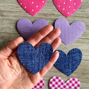 May include: Assortment of fabric heart cutouts in various colors and patterns. Includes pink polka dot, purple, denim blue, and pink gingham hearts. A hand holds a denim blue heart, with others scattered on a wooden surface.