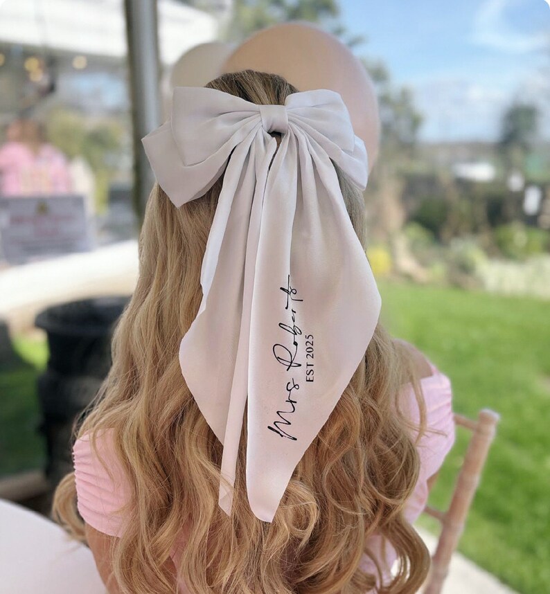 May include: White satin hair scarf with a large bow and the text "Mrs. Roberts Est 2023" printed in black.