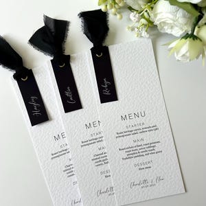 May include: Wedding menu cards with black ribbon tags. Each card displays a menu with starter, main, and dessert options. The cards are white with black text and are decorated with white flowers.