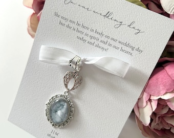 May include: A white card with the text "On our wedding day" and "She may not be here in body on our wedding day but she is here in spirit and in our hearts, today and always!"  A silver charm with a photo and angel wings is attached to a white ribbon.