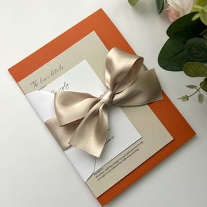 Pode incluir: Um cart&atilde;o de convite dobrado com um cart&atilde;o interior bege e um cart&atilde;o exterior laranja. Uma fita de cetim bege est&aacute; amarrada em um la&ccedil;o na frente do cart&atilde;o. O texto no cart&atilde;o diz "The finer details" e "RSVP".