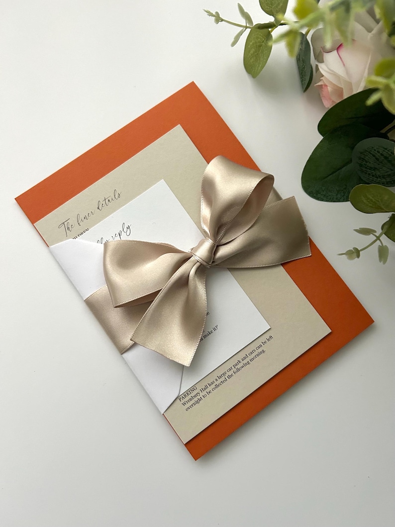 Pode incluir: Um cart&atilde;o de convite dobrado com um cart&atilde;o interior bege e um cart&atilde;o exterior laranja. Uma fita de cetim bege est&aacute; amarrada em um la&ccedil;o na frente do cart&atilde;o. O texto no cart&atilde;o diz "The finer details" e "RSVP".