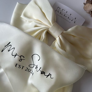 May include: A white satin bow with a white satin ribbon and the text "Mrs. Swan EST 2024" printed on it. The bow is on a white satin background.