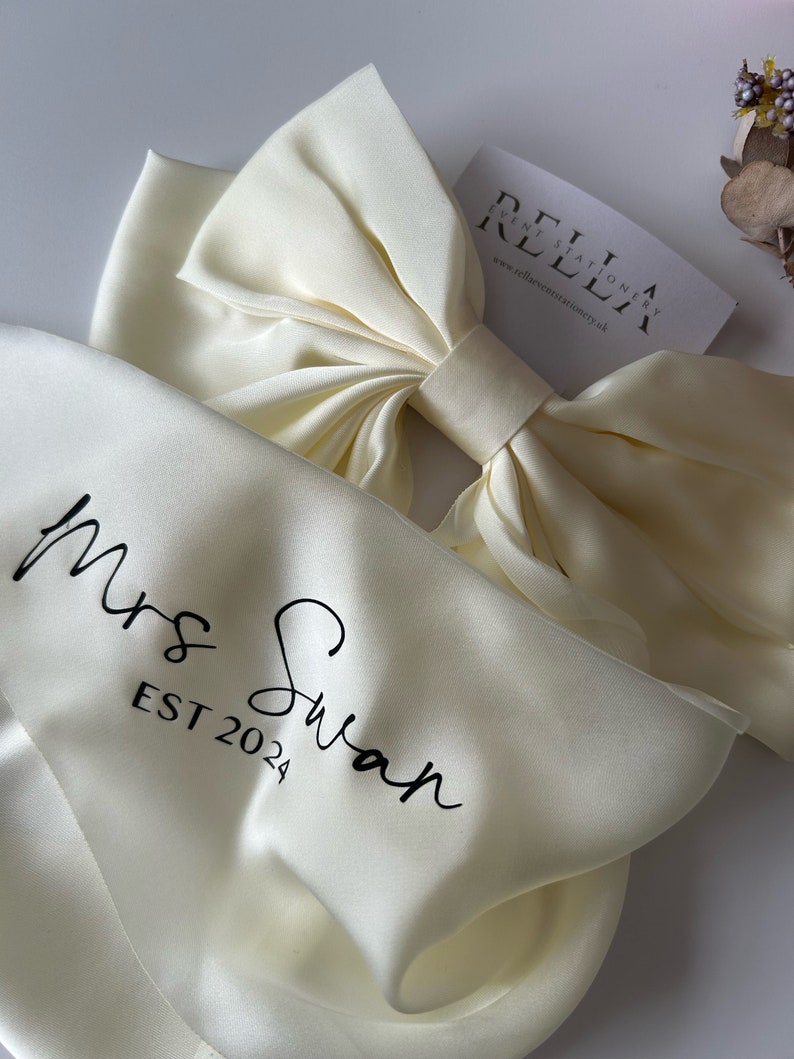 May include: A white satin bow with a white satin ribbon and the text "Mrs. Swan EST 2024" printed on it. The bow is on a white satin background.