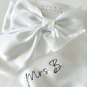 May include: White satin ribbon with a large bow and the text "Mrs. B" written in black calligraphy.