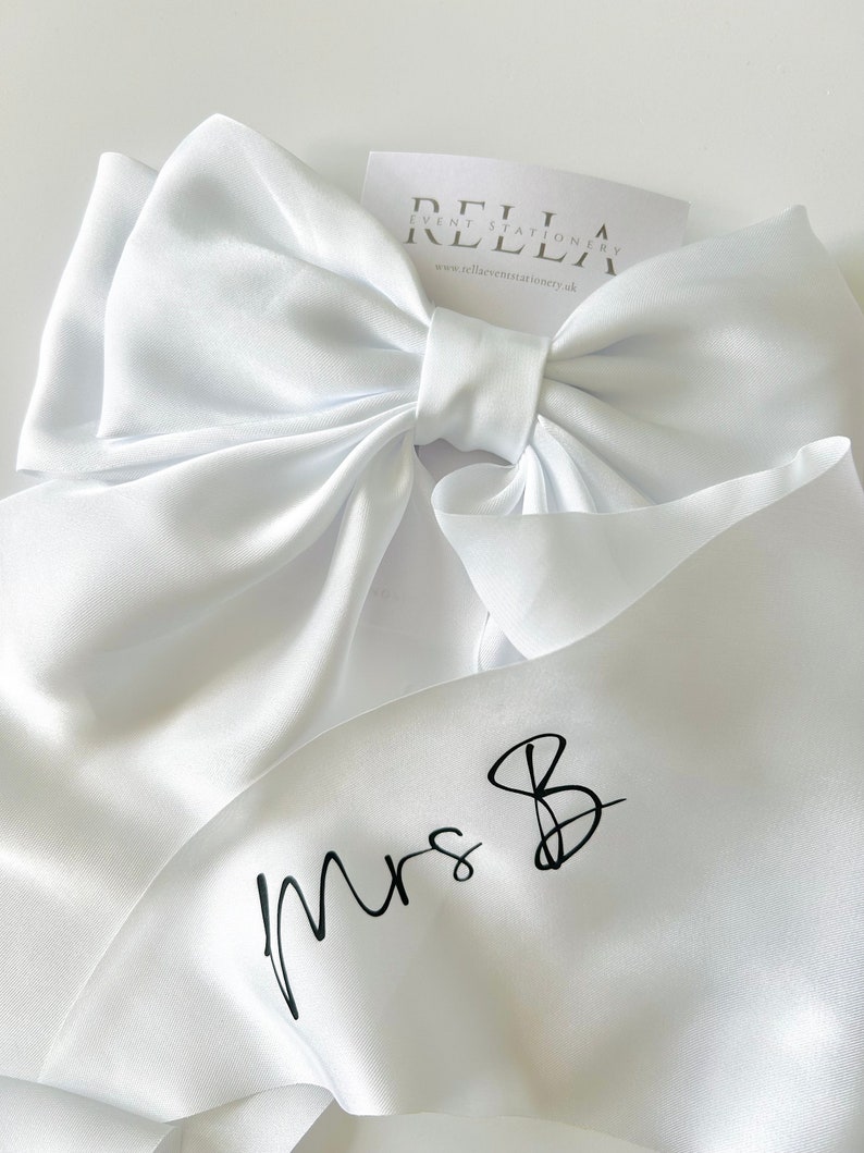 May include: White satin ribbon with a large bow and the text "Mrs. B" written in black calligraphy.