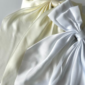 May include: Two large satin bows, one white and one ivory, are arranged on a white surface. The bows are made of soft, flowing fabric and have a classic, elegant design.