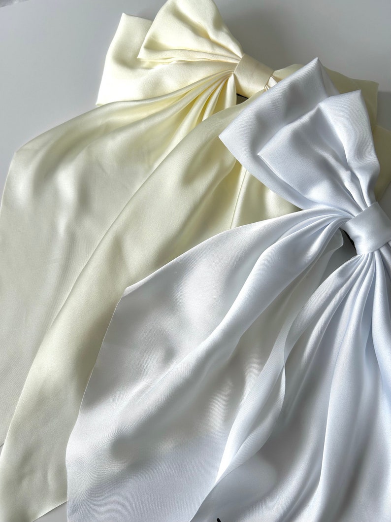 May include: Two large satin bows, one white and one ivory, are arranged on a white surface. The bows are made of soft, flowing fabric and have a classic, elegant design.