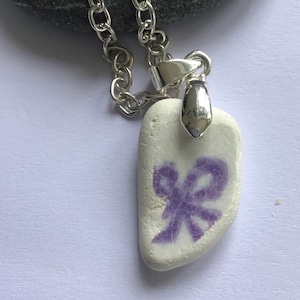 Könnte beinhalten: Eine silberne Kettenkette mit einem weißen Steinanhänger mit einem violetten Schleifendesign.