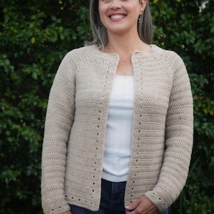 May include: A beige, long-sleeved, open-front crochet cardigan featuring a round neckline and a subtle, repeating vertical stitch pattern along the front edges and cuffs. The garment is worn over a white top and blue jeans.