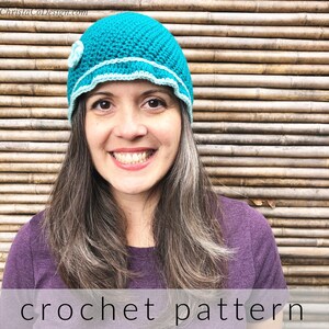 Puede incluir: Un gorro de crochet azul turquesa con un borde de crochet blanco y un acento de flor blanca. El gorro lo lleva una persona con el pelo largo y castaño. El fondo es una pared de bambú marrón.