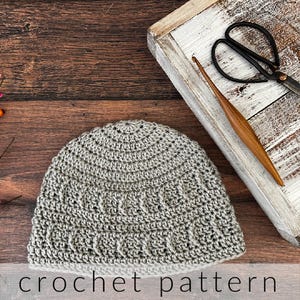 Puede incluir: Un gorro de ganchillo gris claro con un patrón de puntos texturizados con crestas verticales. Junto al gorro hay unas tijeras de metal negro y un ganchillo de madera, sobre una superficie de madera con el texto crochet pattern.