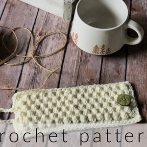 May include: A crocheted cream-colored mug cozy with a button closure. The cozy is lying on a wooden surface with a ball of twine and a mug with a tree design.