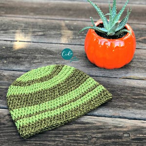 May include: A green and brown striped crocheted beanie hat on a wooden surface. The hat is made with a textured stitch and has a curved brim.