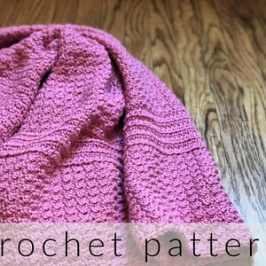 May include: A pink crocheted blanket with a textured stitch pattern. The blanket is folded in half and lying on a wooden surface. The text "crochet pattern" is printed below the blanket.