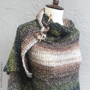 May include: A green, brown, and white ombre knitted shawl with a sparkly finish. The shawl is draped over a mannequin and has a wooden knitting needle pinned through it.