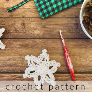 May include: A white crocheted snowflake with a red crochet hook on a wooden background. The background also includes a green and black plaid fabric and a bowl with pine cones.