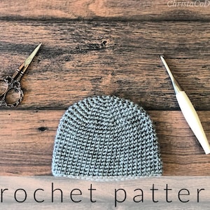 Puede incluir: Un gorro de crochet gris con un borde blanco, unas tijeras y un ganchillo sobre una superficie de madera. patrón de crochet