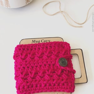 Op de afbeelding: Een roze gehaakte mokwarmer met een knoopsluiting. De mokwarmer zit op een bruin kartonnen kaartje met de tekst "Mug Cozy" en "handmade with love".