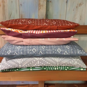 May include: A stack of six rectangular pillows with various patterns and colors. The top pillow is rust-colored with a diamond pattern. Other pillows include pink, blue, and green with geometric designs. The pillows are stacked on a wooden bench.