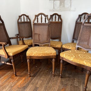 May include: A set of six vintage wooden chairs with cane backs and patterned yellow seat cushions. The chairs have a dark wood finish and ornate detailing, with two armchairs and four side chairs.
