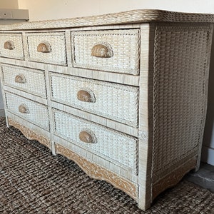 May include: A white wicker dresser with nine drawers and decorative wooden handles. The dresser has a curved top and base with a woven texture. The handles are a light brown color. The dresser is placed on a brown woven rug.