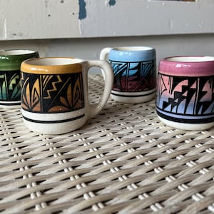 May include: Four small ceramic mugs with white handles and bases. Each mug features a different color scheme: green, yellow, blue, and pink. The mugs have a black band with a geometric design.