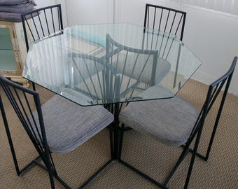 May include: A dining set featuring a hexagonal glass tabletop supported by a black metal frame. Four matching chairs are included, each constructed with a black metal frame and a seat upholstered in a textured gray fabric with a subtle pattern.