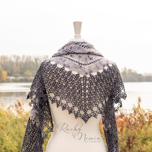 May include: A gray and white crocheted shawl with a lacy design. The shawl is draped over a white mannequin against a backdrop of a lake and fall foliage. The text "Rachy Newin Designs" is visible on the mannequin.