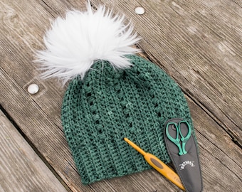 May include: A green crochet hat with a white pom pom, a crochet hook, and a pair of scissors in a black case. The hat is lying on a wooden surface.